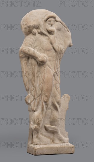 Figurine of Hermes Cryophorus, Statuette of Hermes Cryophorus striding to the right, carrying a goat on his back. His legs are decorated with wings. His face is framed by stubble and a pointed beard. With his left hand, he holds the goat's hind leg under his chin. Depicted in a manner characteristic of the Archaic period: legs in profile, torso and head frontally. The god wears a himation draped like a shawl over his shoulders, ends falling down the sides, and a hairstyle with curls held above his forehead. Hermes strides on an altar, legs depicted in relief against its background. In his bent left hand he holds a caduceus. Set on a small rectangular base. Date: 490–460 BC previously dated 500–490 BC. Place of origin: Boeotia. Type: figurine. Dimensions: height 19.5 cm, width 9 cm, depth 4.5 cm. Weight: 0.28 kg., A pale beige stone sculpture of a robed standing figure shown against a solid gray background, with a rounded hood covering the head and a hand raised toward the face, draped ...