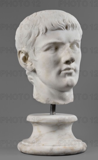Portrait of Drusus the Younger, unknown Roman workshop, male portraits, Roman sculpture, male heads, sketches of male heads, 23–37, Castra Praetoria, Rome, sculpture; height 27 cm, width 20 cm, depth 23 cm, weight 19.5 kg, A white marble sculpted head of a young person with short, textured hair rests on a white marble pedestal connected by a metal rod; the face shows carved eyes, a straight nose, and closed lips with subtle surface blemishes and slight discoloration, and the background is a solid gray color with overall tones of white, gray, and faint beige.