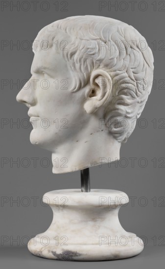 Portrait of Drusus the Younger, unknown Roman workshop, male portraits, Roman sculpture, male head, sketch of male head, 23–37, Castra Praetoria, Rome, sculpture; height 27 cm, width 20 cm, depth 23 cm, weight 19.5 kg, A white marble sculpted head of a man shown in profile on a white marble pedestal against a gray background, with carved short wavy hair, defined facial features including a prominent nose, ear, lips, and chin, subtle surface discolorations and small darker spots on the marble, and a metal rod connecting the head to the rounded pedestal.