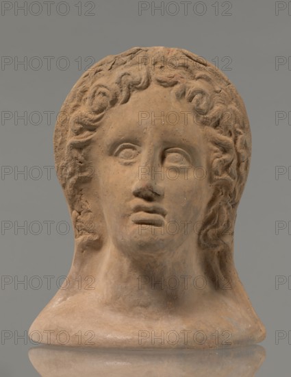 Votive female head, female head wearing a wreath, from under which lush locks of hair curl down the sides of the face to the neck. High forehead, large eyes, deep-set in relation to the nose, slightly convex with developed eyelids and eyeballs, pupils marked. Small straight nose, narrow slightly parted mouth with fleshy lips. Strong chin, slightly protruding. Slim cheeks giving the face a triangular outline. 3rd–2nd century BC, Etruria, votive. Height 26 cm, width 21 cm, depth 13 cm, weight 2 kg., A sculpted head made of a warm brown clay-like material sits against a plain grey background, showing a smooth neck and shoulders and a face with defined brows, a straight nose, full lips, and stylized curly hair framing the forehead and temples; the surface displays subtle variations of lighter and darker brown with small pale flecks and slightly rough texture in areas, and a faint reflected patch of similar brown beneath the base.