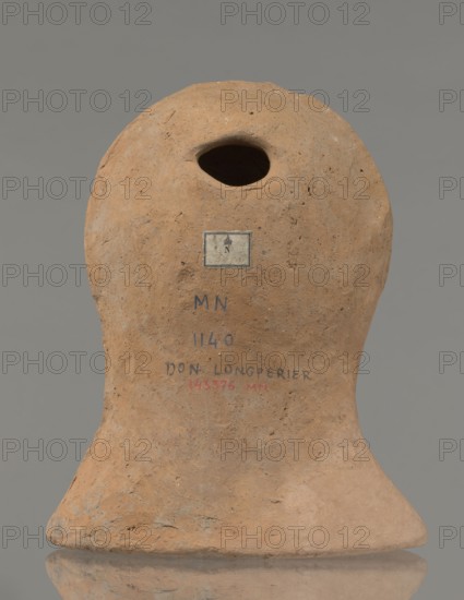Votive female head, Female head wearing a wreath, from under which lush locks of hair curl down the sides of the face to the neck. High forehead, large eyes, deep-set in relation to the nose, slightly convex with developed eyelids and eyeballs, with pupils marked. Small straight nose, narrow slightly parted mouth with fleshy lips. Strong chin slightly protruding forward; slim cheeks giving the face a triangular outline. 3rd–2nd century BC, Etruria; votive. Height 26 cm, width 21 cm, depth 13 cm, weight 2 kg., A rounded earthen object with an irregular oval opening near the top sits against a neutral gray background; its surface is rough and pitted with a natural clay brown color and areas of slightly darker brown and pale tan; a small rectangular label is affixed below the opening and there are traces of dark blue and muted red markings on the front surface; the base flares outward into a slightly uneven foot with similar brown and tan tones and the object casts a subtle reflection on ...