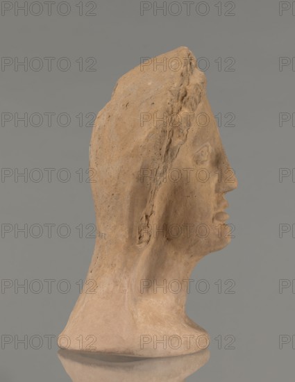 Votive female head, Female head wearing a wreath, from under which lush locks of hair curl down the sides of the face to the neck. High forehead, large eyes, deep-set in relation to the nose, slightly convex with developed eyelids and eyeballs, pupils marked. Small straight nose; narrow, slightly parted mouth with fleshy lips. Strong chin, slightly protruding; slim cheeks giving the face a triangular outline. 3rd–2nd century BC, Etruria; votive. Height 26 cm, width 21 cm, depth 13 cm, weight 2 kg., A sculpted head in profile made of a warm tan clay-like material with visible rough texture and small speckles, showing a defined nose, lips, chin, ear, and a hooded or draped form over the skull; the surface includes areas of smoother and rougher finish and a few small chips or worn spots, all set against a uniform gray background with a faint reflection beneath the base.
