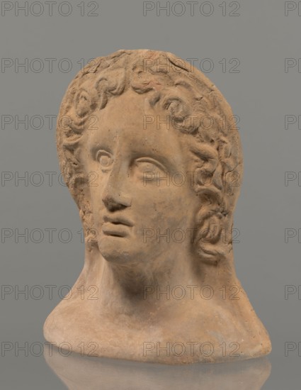 Votive female head, female head wearing a wreath, from under which lush locks of hair curl down the sides of the face to the neck. High forehead, large eyes, deep-set in relation to the nose, slightly convex with developed eyelids and eyeballs, in which the pupils are marked. The nose is small and straight, the mouth is narrow, slightly parted, with fleshy lips. Strong chin, slightly protruding forward. Slim cheeks, giving the face a triangular outline. 3rd–2nd century BC, Etruria, votive. Height 26 cm, width 21 cm, depth 13 cm, weight 2 kg., A sculpted head made of a terracotta-colored material sits against a gray background, showing a stylized face with carved eyes, a straight nose, and closed lips. The hair is rendered in wavy, curled locks that frame the forehead and sides of the face, with textured, slightly rough surfaces and subtle variations in the terracotta tone. The neck and base are visible, with a rounded collar-like bottom and similar earthy coloration, and there are area...