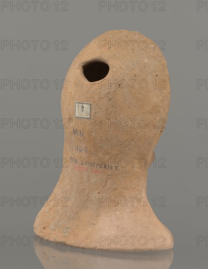 Votive female head, female head wearing a wreath, from under which lush locks of hair curl down the sides of the face to the neck. High forehead, large eyes, deep-set in relation to the nose, slightly convex with developed eyelids and eyeballs with marked pupils. Small straight nose; narrow, slightly parted mouth with fleshy lips. Strong chin, slightly protruding; slim cheeks giving the face a triangular outline. 3rd–2nd century BC, Etruria; votive. Height 26 cm, width 21 cm, depth 13 cm, weight 2 kg., A roughly textured, irregularly shaped clay object in shades of tan and light brown stands against a plain gray background; the object has a rounded top with a deep, dark hollow opening, a sloping curved body with rough surface variations, and a wider base that flares outward and rests on a reflective surface, showing subtle shadows and reflections; there are small darker speckles and patches on the clay, and a few areas of slightly lighter beige where the surface appears smoother.