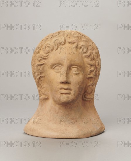 Votive female head. Female head wearing a wreath, from under which lush locks of hair curl down the sides of the face to the neck. High forehead, large eyes deep-set in relation to the nose, slightly convex with developed eyelids and eyeballs, pupils marked. Small straight nose; narrow, slightly parted mouth with fleshy lips. Strong chin, slightly protruding; slim cheeks giving the face a triangular outline. 3rd–2nd century BC, Etruria; votive. Height 26 cm, width 21 cm, depth 13 cm, weight 2 kg., A sculpted head and upper neck made of a warm tan clay sits against a light gray background, showing a face with stylized curly hair, rounded eyes, a straight nose, and closed lips; the surface has subtle variations in tan and ochre with areas of darker brownish spots and faint grayish patches, and the base flares outward into a smooth, rounded pedestal.