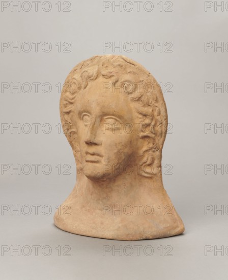 Votive female head;, female head wearing a wreath, from under which lush locks of hair curl down the sides of the face to the neck. High forehead, large eyes, deep-set in relation to the nose, slightly convex with developed eyelids and eyeballs, pupils marked. Small straight nose; narrow, slightly parted mouth with fleshy lips. Strong chin, slightly protruding; slim cheeks giving the face a triangular outline. Etruria, 3rd–2nd century BC; votive. Height 26 cm, width 21 cm, depth 13 cm, weight 2 kg., A single sculpted head made of a warm tan clay sits against a pale gray background, showing a stylized face with softly carved features and curly hair that frames the forehead and sides; the surface displays slight rough texture and subtle variations in the tan color with areas of lighter and darker tan and faint traces of muted gray, and the base flares outward resting on the flat gray surface.
