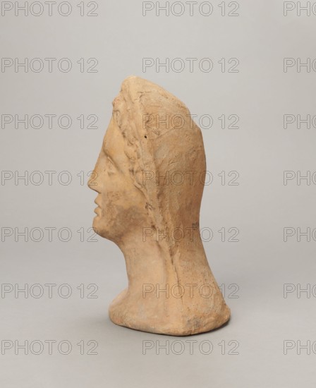 Votive female head, Female head wearing a wreath, from under which lush locks of hair curl down the sides of the face to the neck. High forehead, large eyes deep-set in relation to the nose, slightly convex with developed eyelids and eyeballs with marked pupils. Small straight nose; narrow, slightly parted mouth with fleshy lips. Strong chin slightly protruding forward; slim cheeks giving the face a triangular outline. 3rd–2nd century BC, Etruria; votive. Height 26 cm, width 21 cm, depth 13 cm, weight 2 kg., A sculpted head turned in profile, made of a warm tan clay with areas of lighter and darker tan and pale beige, showing a smooth forehead, prominent nose, closed lips, and a defined chin and neck; the head is covered by a textured hood or headdress with braided detailing along the side, and the base of the piece flares slightly and rests on a neutral pale gray surface against a soft light gray background.