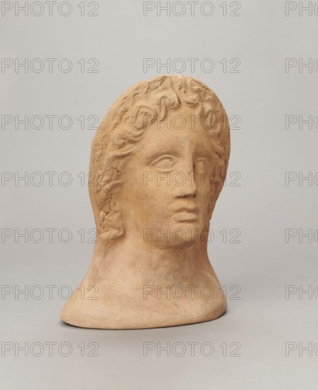 Votive female head, Female head wearing a wreath, from under which lush locks of hair curl down the sides of the face to the neck. High forehead, large eyes, deep-set in relation to the nose, slightly convex with developed eyelids and eyeballs, in which the pupils are marked. The nose is small and straight, the mouth is narrow, slightly parted, with fleshy lips. Strong chin, slightly protruding forward. Slim cheeks, giving the face a triangular outline. 3rd/2nd century BC, Etruria; votive. Height 26 cm, width 21 cm, depth 13 cm, weight 2 kg., A sculpted head in a warm tan clay color sits against a neutral light gray background, showing a smooth neck and base and a face with gently modeled features and wavy hair carved around the forehead and sides; the expression is neutral with slightly parted lips and softly defined eyelids, and the surface shows subtle texture and variations in the tan tones.