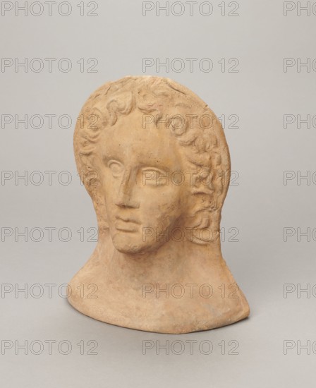 Votive female head, female head wearing a wreath, from under which lush locks of hair curl down the sides of the face to the neck. High forehead, large eyes set deep in relation to the nose, slightly convex with developed eyelids and eyeballs, pupils marked. Small straight nose; narrow, slightly parted mouth with fleshy lips. Strong chin, slightly protruding forward; slim cheeks giving the face a triangular outline. 3rd–2nd century BC, Etruria; votive. Height 26 cm, width 21 cm, depth 13 cm, weight 2 kg., A terracotta bust of a human head and neck sits against a plain light gray background, showing a stylized face with wavy hair framing the forehead and sides; the sculpture's surface is a warm earthy tan with subtle variations of darker and lighter brown, and there are hints of muted orange and pale gray in worn areas, while the background remains mostly pale gray.