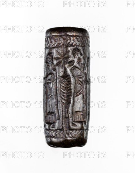 Cylinder seal, unknown Neo-Assyrian workshop. Seal in the so-called linear style with a representation of a religious-magical scene of adoration of the holy tree in the form of a stylised pomegranate bush with a winged solar disc shown above. On both sides of the bush stand human figures in profile, dressed in identical long robes with fringes at the bottom. On the left is a priest, wearing a hat with a ribbon; on the right a king and a courtier, both without headgear. In the field behind the figures are symbols: a palm leaf and an eight-pointed star. The entire image is framed at the top and bottom with a herringbone ornament. Iconography: pomegranate bushes, winged discs, stars. Linear style. 9th–8th century BC; Neo-Assyrian period. Place of origin/ manufacture: Assyria, Northern Mesopotamia; possible finding places: Mesopotamia, Turkey, Syria, Iraq. Seal; height 3.5 cm, width 1 cm., A small cylindrical carved object shown upright against a white background, with a dark brown to blac...