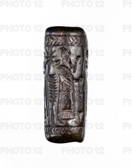 Cylinder seal, unknown Neo-Assyrian workshop. Seal in the so-called linear style with a representation of a religious-magical scene of adoration of the holy tree in the form of a stylised pomegranate bush with a winged solar disc shown above. On both sides of the bush stand human figures in profile, dressed in identical long robes with fringes at the bottom. On the left is a priest, wearing a hat with a ribbon; on the right a king and a courtier, both without headgear. In the field behind the figures are symbols: a palm leaf and an eight-pointed star. The entire image is framed at the top and bottom with a herringbone ornament. Iconography: pomegranate bushes, winged discs, stars. Linear style. 9th–8th century BC; Neo-Assyrian period, Northern Mesopotamia Mesopotamia/Assyria. Materials/type: seal cylinder seal. Dimensions: height 3.5 cm, width 1 cm. Provenance: Turkey, Syria, Iraq findspots/regions., A small, vertically oriented carved object in dark brown and black tones with a slight...