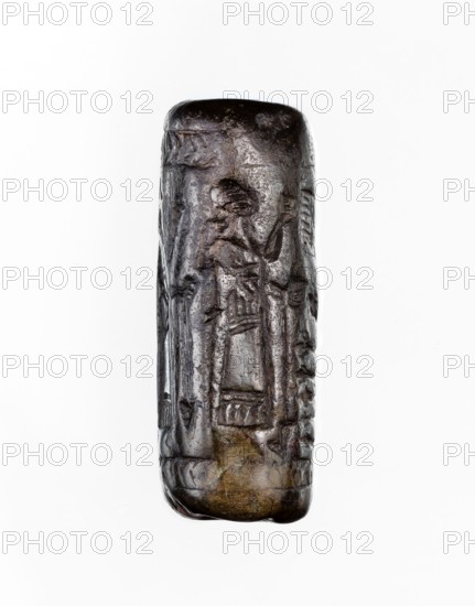 Cylinder seal, unknown Neo-Assyrian workshop. Seal in the so-called linear style with a representation of a religious-magical scene of adoration of the holy tree in the form of a stylised pomegranate bush with a winged solar disc shown above. On both sides of the bush stand human figures in profile, dressed in identical long robes with fringes at the bottom. On the left is a priest, wearing a hat with a ribbon, on the right a king and a courtier, both without headgear. In the field behind the figures are symbols: a palm leaf and an eight-pointed star. The entire image is framed at the top and bottom with a herringbone ornament. Iconography: pomegranate bushes, winged disc, stars. Linear style. Religious scenes. 9th–8th century BC; Neo-Assyrian period, Northern Mesopotamia place of manufacture; Assyria/Mesopotamia/Iraq/Turkey/Syria origins/places of finding. Cylinder seal; height 3.5 cm, width 1 cm., A vertically oriented cylindrical object with an uneven, carved surface showing a few s...
