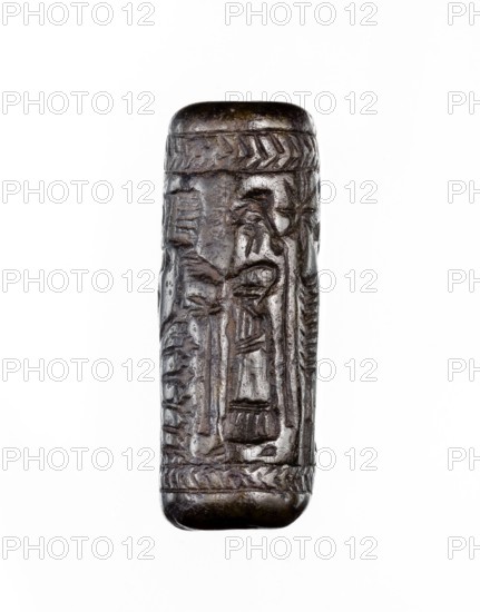 Cylinder seal, unknown Neo-Assyrian workshop. Seal in the so-called linear style with a representation of a religious-magical scene of adoration of the holy tree in the form of a stylised pomegranate bush with a winged solar disc shown above. On both sides of the bush stand human figures in profile, dressed in identical long robes with fringes at the bottom. On the left is a priest, wearing a hat with a ribbon; on the right a king and a courtier, both without headgear. In the field behind the figures are symbols: a palm leaf and an eight-pointed star. The entire image is framed at the top and bottom with a herringbone ornament. Iconography: pomegranate bushes, winged discs, stars. Linear style. 9th–8th century BC; Neo-Assyrian period, Northern Mesopotamia Assyria. Material: seal. Height 3.5 cm; width 1 cm., A small cylindrical carved object with rounded ends, shown against a white background, made of dark brown material with lighter brown highlights; the surface bears carved linear and...