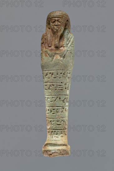Ushabti, Mummy-shaped figurine, funerary shabti, 664–332 BC Late Period, Egypt. Standing on a base, supported by a rear pillar. Arms crossed on the chest with only the hands protruding from under the shroud, holding hoes in both hands. A basket is slung on a single strap over the left shoulder on the back. Engraved tools. Wears a three-part wig styled in narrow strands. Long, tapering beard braided and curled at the bottom. Square face with almond-shaped eyes, pronounced eyebrows, a wide nose, and prominent lips in a slight grimace. Engraved inscription arranged in six lines around the figurine; text illegible. Height 15.5 cm., A small elongated figurine with a humanoid head and a wrapped body, shown against a plain gray background, featuring a weathered surface with shades of green, turquoise, brown, and tan; the head has stylized facial features and a beard, the upper portion shows draped elements and a textured surface, and the lower portion tapers to a rectangular base with surface...