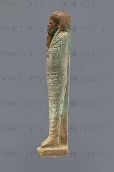 Ushabti, mummy-shaped figurine, funerary gift, hoes. Mummy-shaped figurine standing on a base, supported by a pillar/pilaster at the back. Arms crossed on the chest, only the hands protrude from under the shroud, placed at the same level, close together, holding hoes in both hands. On the back, a basket is slung on a single string over the left shoulder. Engraved tools. On the head, a three-part wig styled in narrow strands. Long tapering beard, curled at the bottom and braided. Square face with almond-shaped eyes, pronounced eyebrows, wide nose, and prominent lips in a slight grimace. Engraved inscription arranged in six lines around the figurine; illegible. 664–332 BC; Late Period, Egypt. Shabti; height 15.5 cm., A slender standing figurine viewed in profile, with a sculpted head and stylized hair in brown tones and a body in greenish-blue and brown patina; the figure appears wrapped in a long garment with subtle raised details and surface texture, showing worn and rough areas, and i...