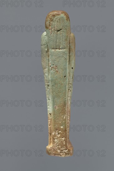 Ushabti, Mummy-shaped figurine, standing on a base, supported by a pillar/pilaster at the back. Arms crossed on the chest, only the hands protrude from under the shroud, placed at the same level, close together. Hoes in both hands. On the back, a basket is slung on a single string over the left shoulder. Engraved tools. On the head, a three-part wig styled in narrow strands. Long, tapering beard curled at the bottom and braided. Square face with almond-shaped eyes, pronounced eyebrows, a wide nose, and prominent lips in a slight grimace. Engraved inscription arranged in six lines around the figurine; illegible text. 664–332 BC; Late Period, Egypt. Shabti; height 15.5 cm., A narrow vertical statuette with a rounded top and flared base, shown against a plain gray background, displaying a weathered surface with a mix of pale green, turquoise, brown, and beige tones; the figure has simplified, elongated form with subtle raised ridges suggesting arms along the sides, pitted and rough areas,...