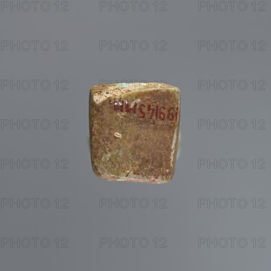 Ushabti, mummy-shaped figurine, funerary gift, hoes. Mummy-shaped figurine standing on a base, supported by a pillar at the back. Arms crossed on the chest with only the hands protruding from under the shroud, placed at the same level, close together, holding hoes in both hands. A basket is slung on a single string over the left shoulder on the back. Engraved tools. Three-part wig styled in narrow strands. Long, tapering beard curled at the bottom and braided. Square face with almond-shaped eyes, pronounced eyebrows, wide nose, and prominent lips in a slight grimace. Engraved inscription arranged in six lines around the figurine text illegible. 664–332 BC, Late Period, Egypt. Shabti; height 15.5 cm., A small, worn rectangular object with rounded corners floats against a plain gray background; its surface shows mottled shades of brown and green with patches of lighter beige and a few small darker spots, and there is a hint of a reddish marking on one side.