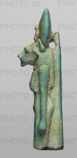 Figurine amulet of the goddess Sachmet lion-headed, possibly Bastet?, wearing a tripartite wig and a long narrow robe, topped with a solar disc with a uraeus. The lower legs are missing; the figure appears to be walking with arms lowered. Back pillars. Late Period, Egypt, 664–332 BC. Material: amulet; height 5.8 cm, width 1.7 cm., A slender vertical figurine in shades of turquoise, teal, pale green, and sandy beige shows a stylized anthropoid profile with an elongated head and pronounced snout, a rounded ear or headpiece, and a visible arm folded along the body; surface texture appears worn and pitted with areas of darker green and small patches of brownish discoloration, and there is a circular hole near the upper rear of the piece.