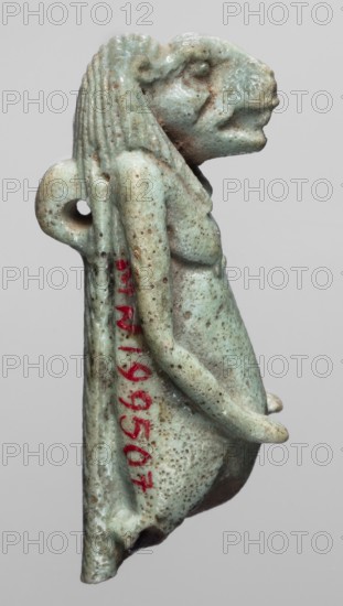 Amulet — Toeris; maker, subject: Toeris Egyptian hippopotamus goddess; Egyptian religion; Late Period, 664–332 BC, Egypt; material: amulet; height 4.6 cm, width 1.5 cm; noted as repossession Radom., A small sculpted figure viewed in profile against a plain light gray background, showing a pale greenish-blue surface with mottled brown spots and areas of darker discoloration; the figure has a humanoid body with a pronounced snout-like face, a textured mane or hair falling down the back, rounded shoulders, and an arm bent along the torso; a loop or hole is visible near the upper back, and there are traces of reddish pigment on the rear side; the surface appears pitted and weathered with a glossy, glazed finish.