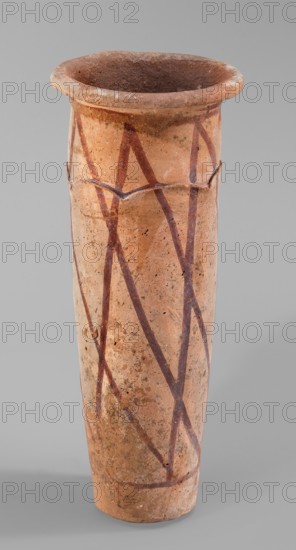 Cylindrical vessel with crosshatched decoration, painted ceramic with engraved decorations and geometric motifs. The vessel has a shape resembling a cylinder, slightly tapering towards the bottom, with a slightly convex bottom and a widely rolled-out rim. A plastic, wavy ornament runs 3/4 of the way up; it is arranged so the upper part of the vessel appears recessed relative to the lower. Surface covered with a light engobe and decorated with a painted diagonal grid. Attributed to unknown author, possibly B. Ruszczyc. c. 3200–c. 3050 BC; Predynastic Period, Naqada III Naqada phase IIIa2, from Abusir el-Melek. Vessel. Height 27.5 cm; width 11 cm; weight 1.028 kg., A tall cylindrical ceramic vessel with a flared rim, showing a worn, textured surface in shades of light brown and tan with darker brown and reddish-brown painted zigzag lines and spots; the interior appears darker brown, and the background is a neutral gray.