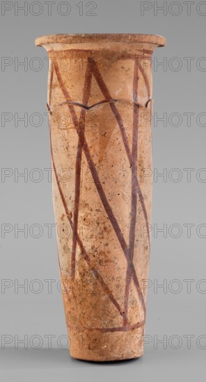 Cylindrical vessel with crosshatched decoration, painted and engraved ceramic, geometric motifs. The vessel has a slightly tapering cylindrical shape with a slightly convex bottom and a widely rolled-out rim. A plastic, wavy ornament runs three-quarters of the way up, arranged so the upper part appears recessed relative to the lower. Surface covered with a light engobe and decorated with a painted diagonal grid. Attributed to B. Ruszczyc, c. 3200–c. 3050 BC; Predynastic Period, Naqada III Naqada phase IIIa2, Abusir el-Melek find site. Vessel; height 27.5 cm, width 11 cm, weight 1.028 kg., A tall cylindrical terracotta vessel with a flared rim, showing an overall beige and light brown clay surface with darker brown and reddish-brown painted diagonal lines forming a zigzag pattern; the vessel surface has areas of darker brown speckling and spots, some rough and worn patches, and a few chips along the rim and body, set against a neutral gray background.