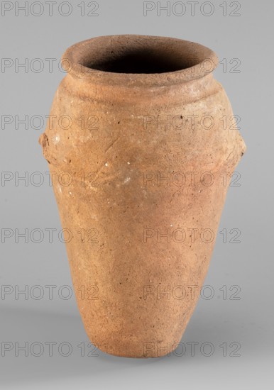 Vessel with wavy handles., Vessel with an elongated belly that widens towards the top. Short, cylindrical neck and rounded, upturned rim. At approx. 2/3 of the height two residual vices resembling an attached roller with a wavy profile are preserved; the vices are placed asymmetrically. Flat bottom. Even surface with scratch marks visible in the upper part. Clay color: light red. c. 3500–3050 BC; Predynastic Period, Naqada II/III Naqada phase IId2–IIIa1. Found at Abusir el-Melek. Vessel for oils and ointments. Egyptian ceramics; height 20.2 cm, width 13.3 cm, weight 0.708 kg., A single large earthenware vessel fills the frame, with a rounded rim opening into a dark interior and a tapered body that narrows toward the base; the surface is rough and textured with small pits and irregularities and shows a few raised areas where clay was added. The visible colors are shades of brown and tan on the vessel, with small white specks scattered across its surface, set against a smooth light gray ...