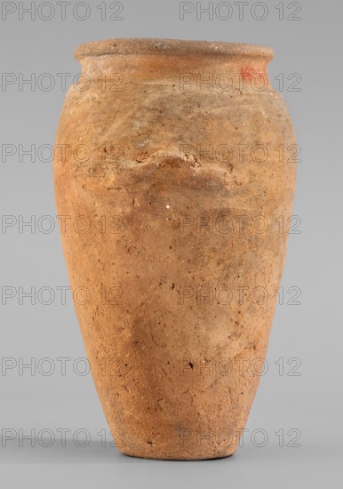 Vessel with wavy handles. Egyptian ceramic vessel for oils and ointments, c. 3500–3050 BC, Predynastic Period, Naqada II/III Naqada phase IId2–IIIa1, found at Abusir el-Melek. Vessel with an elongated belly that widens toward the top; short cylindrical neck; rounded, upturned rim. At approx. two-thirds of the height two residual vices resembling an attached roller with a wavy profile are preserved, placed asymmetrically. Flat bottom. Even surface with scratch marks visible in the upper part. Clay color light red. Height 20.2 cm, width 13.3 cm, weight 0.708 kg., A tall, tapered clay vessel occupies the frame, showing a rough, grainy surface with visible cracks and small pits; its body is predominantly warm terracotta with areas of darker brown and lighter beige variations, and there are tiny white flecks scattered across the surface; the rim is slightly flared and shows a more concentrated band of lighter brown, while the background is a smooth, neutral gray.