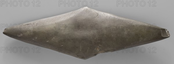 Rhomboid palette. Palette in the shape of an elongated rhombus; possible whetstone. Ceremonial/cosmetic object, geometric palette, toiletry item. 1st half of the 4th millennium BC; Predynastic Period Naqada I phase, Egypt. Height 24 cm, width 8 cm, weight 0.316 kg., An elongated, symmetrical object with pointed ends resting against a plain background, showing a smooth, slightly uneven surface with visible scratches and small pits; the object appears predominantly dark gray to charcoal with areas of lighter gray highlights and a few tiny spots of brownish discoloration, while the background is a uniform light gray.