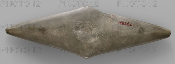 Rhomboid palette. Palette in the shape of an elongated rhombus; whetstone? Ceremonial/cosmetic palette and toiletry item made of stone, geometric palette. 1st half of the 4th millennium BC; Predynastic Period Naqada I phase, Egypt. Height 24 cm; width 8 cm; weight 0.316 kg., A long, symmetrical, elongated object with pointed ends resting on a flat neutral background, showing a smooth, slightly irregular surface with visible scratches and worn patches; the object is primarily gray-green with areas of lighter gray and muted brownish spots, and there are small darker smudges and pale scuffed areas across its surface.