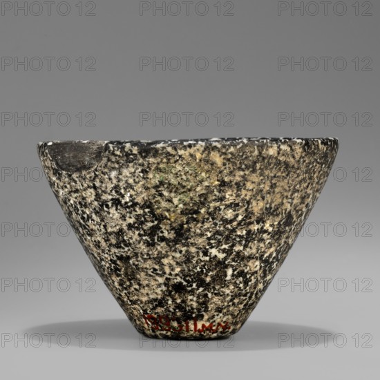 Bowl with high walls, widening towards the top. The rim is rounded and slightly beveled inwards. The bottom is small and flat. Stone vessel, ca. 3100–ca. 2686 BC Early Dynastic Period, Egypt., Vessel; height 6.7 cm, width 4.9 cm, weight 0.30 kg., A roughly conical stone vessel with a wide rim tapering to a narrower base sits against a neutral gray background; the surface shows a speckled pattern of black, white, beige, and small areas of green, with a slightly chipped or worn section near the rim and a soft shadow beneath the object.
