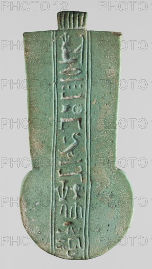 Amulet - weight of the menit necklace of the priest Achpet, menit, Achpet ca. 664–404 BC, hieroglyphs, priests, necklace weight with a column of hieroglyphic inscription through the center, 664–332 BC; Late Period, Egypt, amulet, height 8.7 cm, width 3.5 cm, An elongated, flattened object with a rounded lower end and a narrower rectangular upper section, shown against a plain pale gray background; the surface is a mottled green with areas of lighter and darker green and small patches of brownish discoloration and pitting; a central vertical recessed panel runs the length of the piece, bordered by shallow grooves, and contains a series of raised shapes and shallow incisions; the top has a short, ribbed projection with vertical grooves and the edges are slightly worn and uneven, revealing some rougher brownish areas.