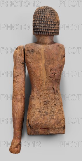 Male statue by Genius, Mieczyslaw 1853–1920. Egyptian sculpture, Old Kingdom, 6th Dynasty, c. 2345–c. 2181 BC. Materials/attributes: grave equipment; wigs; slender torso, flat at hip level; thick, short neck; round head; almond-shaped eyes with marked irises; broad nose; thick mouth; left arm extended downwards with hand clenched into a fist. Dimensions: height 56 cm, width 18 cm, depth 10.5 cm., A carved wooden figure shown from the back with a textured, grid-patterned rounded head in dark brown and black tones, a torso and detached arm in various shades of brown and tan with visible cracks and rough surface texture, and a pale gray background.