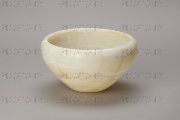 Bowl, Large stone bowl with a small flat bottom, slightly flared side walls bent inwards at the top and a straight rim. Old Kingdom, ca. 2686–2181 BC. Found at Abu Rawash. Vessel; height 14 cm, width 23 cm, weight 1.35 kg., A smooth rounded bowl sits on a flat surface, showing a glossy stone-like texture with subtle veining and irregular patching; the bowl displays shades of creamy white, pale yellow, warm beige, and hints of light gray, with soft highlights and gentle shadows on its interior and exterior surfaces, and a neutral gray background and surface beneath it.