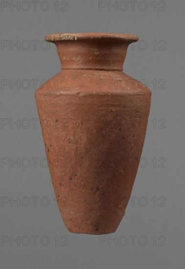 Vessel, Egyptian pottery, ceramics, clay vessel, conical-shaped vessel, widest at approximately 3/4 of its height. Quite wide, almost horizontal shoulders slightly sloping. Wide, short, funnel-shaped neck, widening towards the spout. Wide spout, flat in the upper part, slightly rounded at the bottom, slightly asymmetrical. Flat bottom, asymmetrical. Surface leveled. Lower part finished to approximately 2/3 of its height with a knife or similar tool. Outer surface and inner surface of the rim and neck covered with a layer of dark red engobe. Vessel color dark red. Vessel complete; surface eroded in many places, numerous clay chips on the rim. Ca. 2686–ca. 2181 BC; Old Kingdom, Edfu, vessel, height 16 cm, diameter 8.6 cm rim, diameter 10.7 cm body, diameter 4.5 cm bottom., A single tall earthenware vessel with a wide flaring rim and a tapered body fills the image against a plain background. The surface of the vessel is textured and worn, showing rough patches and small pitted areas. Its ...