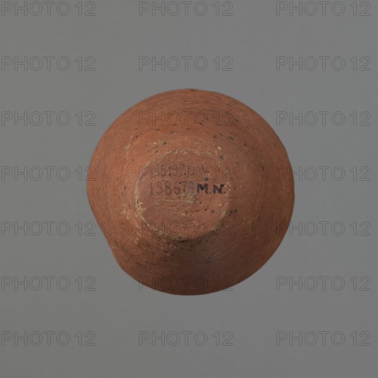 Vessel, Egyptian pottery, ceramics, clay vessel. Conical-shaped vessel, widest at approximately 3/4 of its height. Quite wide, almost horizontal shoulders slightly sloping. Wide, short, funnel-shaped neck, widening towards the spout. Wide spout, flat in the upper part, slightly rounded at the bottom, slightly asymmetrical. Flat, asymmetrical bottom. Surface leveled. Lower part finished to approximately 2/3 of its height with a knife or similar tool. Outer surface and inner surface of the rim and neck covered with a layer of dark red engobe. Vessel color dark red. Vessel complete; surface eroded in many places, numerous clay chips on the rim. Old Kingdom, ca. 2686–2181 BC; provenance Edfu. Vessel height 16 cm; diameter 8.6 cm rim, 10.7 cm body, 4.5 cm bottom., A rounded terracotta object with a rough, speckled surface in shades of reddish-brown and warm orange sits against a flat gray background, showing a recessed circular base area with worn edges and traces of a darker brown patina; ...