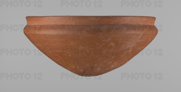Medum type bowl, Medum type bowl with carination under the rim. Rim diameter smaller than the diameter of the shoulders. Rounded bottom. Straight, slightly flaring rim. Vessel made on the wheel of Nile clay, surface leveled. The bottom and lower part of the vessel finished to about half its height with a knife or similar tool. The surface of the vessel is covered with a layer of engobe . Vessel colour – red. c. 2686–c. 2181 BC; Old Kingdom, Edfu. Vessel; height 9 cm; diameter 21.1 cm rim, 21.5 cm shoulders., A large terracotta-colored ceramic bowl is shown from a side view against a neutral gray background, with a rounded body that tapers to a pointed base and a slightly flared rim. The bowl's surface shows variations of reddish-brown and orange hues with areas of lighter tan and pale spots, plus visible scuffs and subtle surface texture. The rim has a darker band of brownish-orange, and there are small dark specks and worn marks scattered across the exterior.