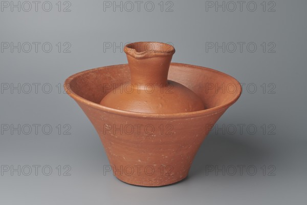Deep bowl. Deep, cup-shaped bowl with a slightly convex bottom and sides arching upwards; straight, flat-topped, flaring rim. Made of Nile clay NB1 ? per Vienna System. Covered with an engobe containing red ochre and highly polished. Vessel color: red. Well-preserved with slight clay damage on rim. Comparative references associate similar types with 4th–6th Dynasties/Old Kingdom contexts. Dimensions: height 15.1 cm; lip diameter 27.6 cm; bottom diameter 11.8 cm; weight 1.3 kg. Maker, likely Egyptian ceramic, c. Old Kingdom period., An orange-brown clay vessel with a wide, flaring rim and a narrower base sits centered on a smooth gray surface against a gray backdrop; the vessel has a rounded inner bulb with a short cylindrical neck and a slightly undulating lip, showing a matte, textured surface with small speckles and patches of lighter wear.