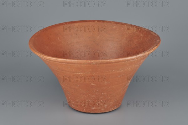 Deep, cup-shaped clay bowl with a slightly convex bottom and sides arching upwards; straight, flat, flaring rim. Made of Nile clay NB1 ? and covered with an engobe mixed with red ochre and thoroughly polished, giving a red color. Well preserved with slight clay damage on the rim. Comparative parallels place it among Old Kingdom types 4th–6th Dynasty; measurements: height 15.1 cm, lip diameter 27.6 cm, bottom diameter 11.8 cm, weight 1.3 kg., A wide, shallow bowl-shaped object with a flared rim sits on a smooth flat surface; it appears made of an earthy terracotta material showing a reddish-brown color with lighter worn patches and small speckles, and the interior and exterior surfaces share the same reddish-brown hue with subtle variations; the background and surface are a cool gray, creating contrast with the pot's warm tones.