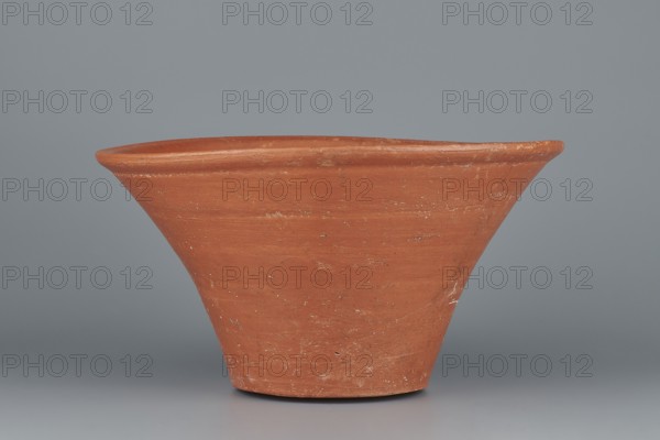 Deep, cup-shaped bowl with a slightly convex bottom and sides arching upwards; straight, flat, flaring rim. Made of Nile clay NB1 ? according to the Vienna System, covered with an engobe containing red ochre and thoroughly polished. Color: red. Well-preserved with slight clay damage on the rim. Comparative attributions: cited parallels to pottery and typologies from the 4th–6th Dynasties / Old Kingdom. Vessel dimensions: height 15.1 cm; lip diameter 27.6 cm; bottom diameter 11.8 cm; weight 1.3 kg., A single terracotta-colored clay vessel with a flared rim and conical body sits against a smooth gray background; the surface of the vessel shows variations in the terracotta hue with lighter and darker patches and some small worn or rough spots, and the scene includes shades of gray in the backdrop and base area.