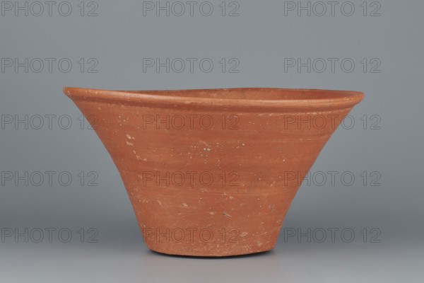 Deep, cup-shaped clay bowl with a slightly convex bottom and sides arching upwards; straight, flat, flaring rim. Made of Nile clay NB1 Vienna System. Covered with an engobe mixed with red ochre and thoroughly polished; red color. Well-preserved with slight clay damage on the rim. Function: vessel. Comparative typology links to Old Kingdom and late 6th Dynasty pottery 4th–6th Dynasty. Dimensions: height 15.1 cm; lip diameter 27.6 cm; bottom diameter 11.8 cm; weight 1.3 kg., A terracotta-colored, conical clay pot with a flared rim sits against a smooth gray background; the pot shows a textured surface with small pale speckles and slight irregularities in the earthy orange-brown clay, and the lighting creates soft shadows beneath and to the sides, highlighting the pot’s rounded lip and tapering base.