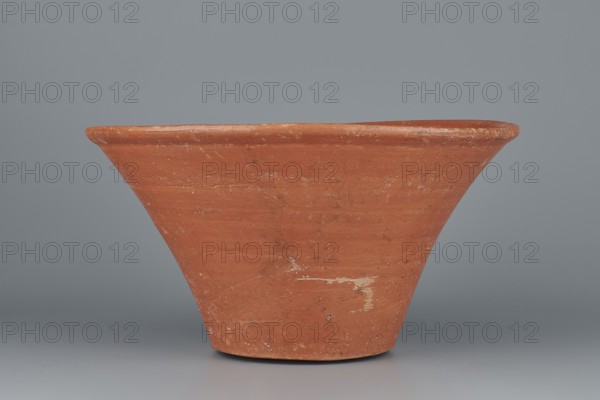 Deep bowl. Deep, cup-shaped bowl with a slightly convex bottom and sides arching upwards; straight, flat, flaring rim. Made of Nile clay NB1, Vienna System. Covered with an engobe mixed with red ochre and thoroughly polished. Vessel color: red. Well-preserved, slight clay damage on the rim. Comparative typology: parallels cited for Old Kingdom and late 6th Dynasty pottery 4th–6th Dynasty. Dimensions: height 15.1 cm; lip diameter 27.6 cm; bottom diameter 11.8 cm; weight 1.3 kg., A wide, shallow clay vessel with a flared rim sits against a smooth backdrop; the vessel is terracotta orange with patches of lighter and darker orange and some worn pale areas, showing rough texture and small blemishes, and the background and surface beneath the vessel are shades of gray.
