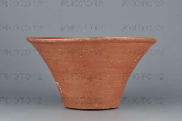 Deep bowl. Cup-shaped clay bowl with a slightly convex bottom and sides arching upwards, straight flaring rim flat on top. Made of Nile clay NB1 ? with an engobe containing red ochre, thoroughly polished. Color: red. Well-preserved with slight clay damage on the rim. Function: vessel. Dimensions: height 15.1 cm; lip diameter 27.6 cm; bottom diameter 11.8 cm; weight 1.3 kg. Attributed date/style: Old Kingdom, 4th–6th Dynasty c. 2494–c. 2181 BC. Comparative parallels: parallels noted with Mycerinus and Giza Necropolis pottery and late 6th-dynasty and Early Bronze Age III types. Maker: unknown., A wide, flared terracotta vessel with a rough, matte surface in shades of orange-brown and reddish-brown, showing lighter beige and white spots and scuffs, set against a smooth, cool gray background; the vessel tapers toward a narrower base and has a slightly rounded rim with visible texture variations.