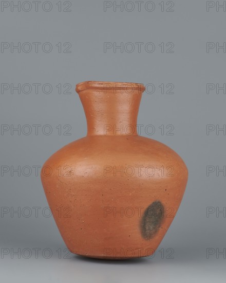 Jug, Medium-sized biconical clay jug with wide, sloping shoulders and a wide neck that is almost cylindrical in its lower section and widens toward the spout neck height ˜ 1/3 of total. Profiled, irregular spout raised and extended on one side forming a small secondary spout; slightly rounded, almost triangular in cross-section, with a millimetre-long indentation on the outer profile. Vessel is widest at about half its height; bottom almost flat very slightly rounded. Made on the wheel from Nile clay NB1 Vienna System. Outer surface leveled, covered with an engobe and polished; inner surface leveled. Color red-brown. Complete, very well preserved. Old Kingdom, 5th–6th Dynasty, c. 2494–c. 2181 BC; find place: Edfu. Vessel. Height 19.9 cm; rim diameter 8.1 cm; bottom diameter 9.5 cm; body diameter 16.7 cm; weight 1.1 kg., A rounded clay vessel in a terracotta orange color with a flared neck and slightly pinched rim sits against a smooth gray background; the surface of the vessel shows a ...