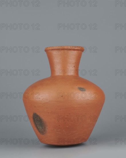 Jug. Medium-sized biconical clay jug with wide, sloping shoulders and a wide neck that is almost cylindrical in its lower section and widens toward the spout, the neck about one-third of the vessel's height. Profiled, irregular spout raised and extended on one side forming a small secondary spout; slightly rounded, almost triangular in cross-section with a millimetre-long indentation on the outer profile. Bottom almost flat, very slightly rounded. Made on the wheel of Nile clay NB1 Vienna System. Outer surface leveled, covered with an engobe and polished; inner surface leveled. Color red-brown. Complete and very well preserved. Old Kingdom, 5th–6th Dynasty, c. 2494–c. 2181 BC, from Edfu. Vessel; height 19.9 cm, rim diameter 8.1 cm, bottom diameter 9.5 cm, body diameter 16.7 cm, weight 1.1 kg. Analogies: T. Rzeuska, Saqqara II; G. Brunton, Qau and Badari., A terracotta-colored pottery vessel with a rounded body that tapers toward a short, flared neck and a slightly rolled rim, showing a...