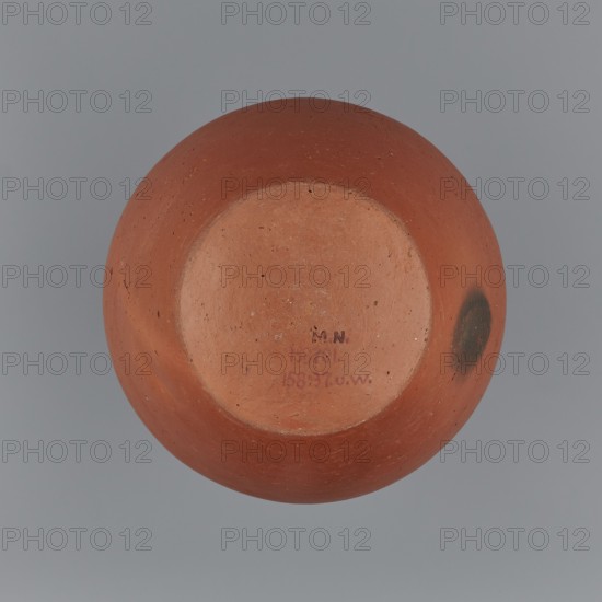 Jug. Medium-sized biconical clay jug with wide, rather sloping shoulders and a neck nearly cylindrical in its lower section, widening toward the spout neck height ˜ 1/3 of vessel. Profiled, irregular spout raised and extended on one side to form a small secondary spout; slightly rounded, almost triangular in cross-section with a ~1 mm indentation on the outer profile. Vessel widest at about half its height; bottom almost flat, very slightly rounded. Made on the wheel from Nile clay NB1 Vienna System. Outer surface leveled, covered with an engobe and polished; inner surface leveled. Red-brown in color. Complete and very well preserved. c. 2494–c. 2181 BC, Old Kingdom, 5th–6th Dynasty, Edfu. Vessel; height 19.9 cm; rim diameter 8.1 cm; bottom diameter 9.5 cm; body diameter 16.7 cm; weight 1.1 kg., A rounded clay object viewed from below showing a circular recessed base and a smooth curved body, with an overall terracotta color and a darker burnished spot on one side; the surface has smal...