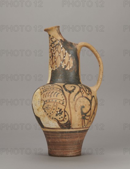 Red-figure oinochoe by the Pennsylvania Torcop Painter, Classified Gallo-Classic Scenography; red-figure ceramic wine vessel depicting mythological scenes and women; 2nd half of the 4th–3rd century BC c. 300–280 BC, to be verified; Etruria place of origin; vessel; height 31.5 cm, width 16.2 cm, depth 15.6 cm, weight 0.94 kg, A ceramic jug with an elongated neck, curved handle, and rounded body standing on a conical foot; the surface shows painted decorative patterns and areas of worn or flaked paint. The visible colors include beige and tan on the clay body, black and dark brown used for painted motifs and a darker area on the neck, white applied in small spots and patches, and reddish-brown bands around the foot. The decoration features flowing curved lines, spiral-like shapes, dotted accents, and a lattice-like pattern on the upper neck, with some areas where the paint is chipped or missing revealing the underlying lighter clay.