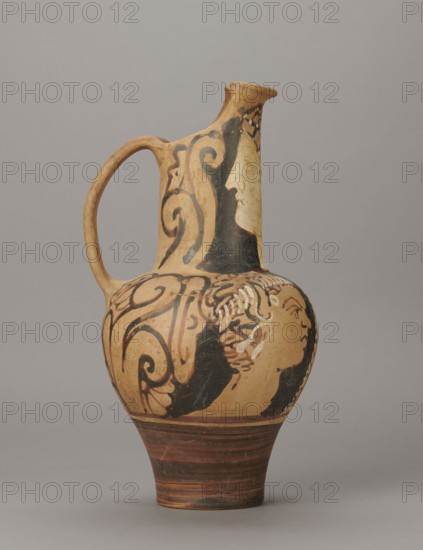 Red-figure oinochoe by the Torcop Painter, Classified Gallo-Classic Scenography; red-figure pottery, wine vessel depicting mythological scenes and women; 2nd half of the 4th–3rd century BC c. 300–280 BC, to be verified; Etruria place of origin; vessel; height 31.5 cm, width 16.2 cm, depth 15.6 cm, weight 0.94 kg, A ceramic jug with a rounded body, narrow neck, and single loop handle, displayed against a neutral gray background; the jug has an overall warm beige base color with areas of black painted decoration forming flowing scroll-like shapes and partial human profiles, and there are accents of darker brown on the lower foot and rim.