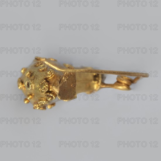Leech fibula, brooch made of gold plate with a swollen ring decorated with filigree rings and central granulation. No sheath. 6th century BC, Etruria. Jewelry fibula. Height 1.7 cm, width 2.1 cm, depth 0.7 cm, weight 0.7 g., A small gold-colored metallic object with a textured spherical end covered in a few raised bumps and nodules, connected to a flat elongated arm and a short irregular block-like section; the object rests against a pale gray background and shows highlights and reflections in shades of bright gold and darker golden tones.