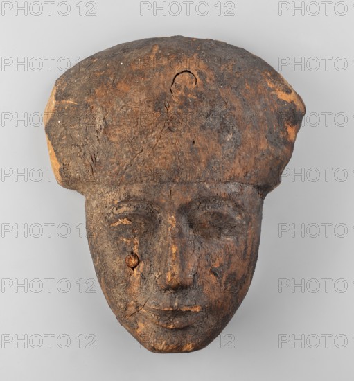 Sarcophagus mask; wig iconographic; sarcophagus fragments; masks, faces; anthropoid sarcophagus mask — face with fragment of a wig above the forehead. Facial features rendered in plastic. Traces of black paint. c. 1069–c. 945 BC; Third Intermediate Period; 21st Dynasty; Deir el-Medina; sarcophagus mask., A worn carved wooden face mask with a rounded headdress, showing a textured surface with areas of dark brown and lighter brown wood, some blackened patches, and a few small pale spots; the face has carved eyes, a nose, and a closed mouth, and there is visible cracking and rough grain throughout the piece.