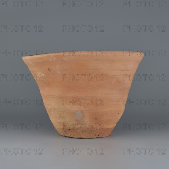 Cake baking mold so-called flower pot for baking bread with a conical shape and a flat bottom. Irregular shape with a straight, flaring spout and an irregular bottom. Wheel-made from Nile clay of type NB2 Vienna System. Surface level; outer part near the bottom and the bottom finished with a knife or similar tool. Hole in the bottom ~6 cm diameter made before firing. Yellowish-red color. Very well preserved. Rim diameter 22.3–25.2 cm; bottom diameter 10.2–11 cm; height 17.4 cm. c. 1550–c. 1069 BC, New Kingdom, Deir el-Medina., A terracotta-colored clay pot with a flared rim sits against a smooth gray background; the pot shows a natural matte texture with areas of lighter and darker orange-brown, some small blemishes and smudges, and a rougher, slightly darker base, while the scene includes shades of gray in the backdrop and surface.